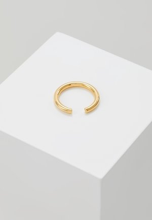 Offener polierter Goldring auf einer weißen kubischen Plattform vor einem hellgrauen Hintergrund dargestellt.