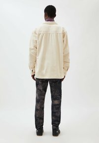 Lichtbeige corduroy overhemdjack met een kraag, lange mouwen en knoopmanchetten; gecombineerd met donker tie-dye broeken en zwarte schoenen.