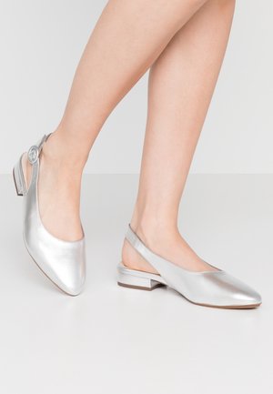 Kobieta nosząca srebrne buty na niskim obcasie typu slingback, stojąca na jasnoszarej powierzchni na tle jednolitego jasnoszarego tła.