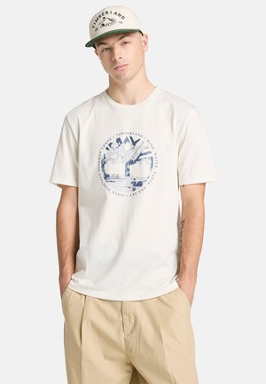 Beżowy bawełniany t-shirt z niebieskim logo graficznym przedstawiającym górę na środku, zestawiony z beżowymi spodniami i jasną czapką.