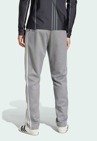 Grå atletiska sweatpants med vita sidoremmar, tillverkade av mjukt tyg, i kombination med vita skor med svarta ränder, sedd bakifrån.