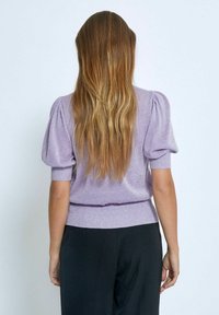 Pull manches courtes lavande avec épaules bouffantes, ourlet côtelé et texture brillante. Porté avec un pantalon noir, affichant une silhouette lisse.