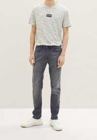 Maglietta grigia a maniche corte con etichetta nera, abbinata a jeans grigio scuro e sneakers bianche. L'outfit presenta una vestibilità casual e un design semplice.