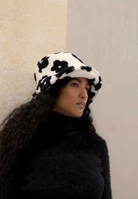 Melnā un baltā floreļa spainītis cepure, kas izgatavota no mīkstas, teksturētas auduma. Tam ir plats, noapaļots maliņš un ērta piegulšana. Valkāta kopā ar melnu augsto kaklu.