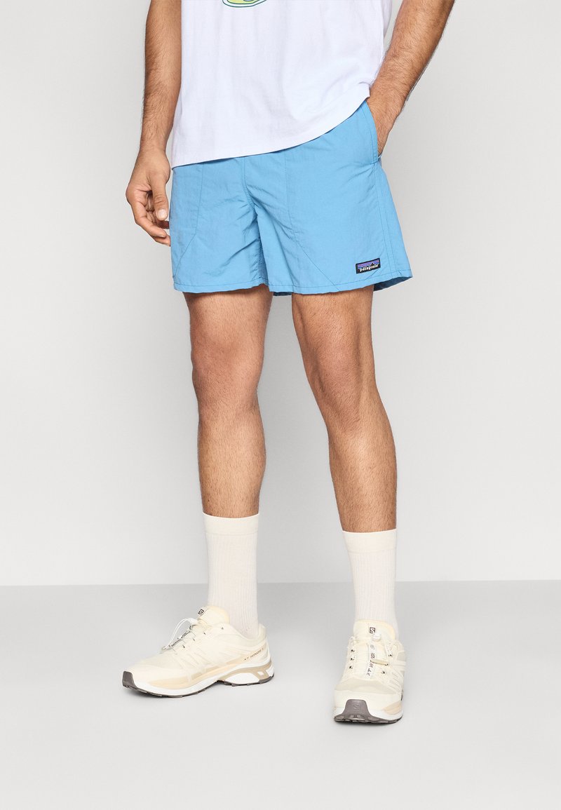 Ανοιχτό μπλε μαγιό σορτς με πλαϊνές τσέπες, κατασκευασμένα από γρήγορο στέγνωμα υλικό. Συνδυάζονται με μπεζ αθλητικά παπούτσια και λευκές κάλτσες κοντές για αντίθεση.