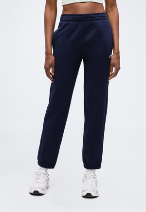 Vrouw draagt marineblauwe joggingbroek met elastische boorden en witte sneakers, staand tegen een effen lichte achtergrond.