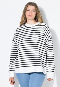 Sudadera a rayas en blanco y negro con mangas largas, que presenta un cuello redondo y un pequeño acento de logo verde en el pecho. Combinada con jeans azules.