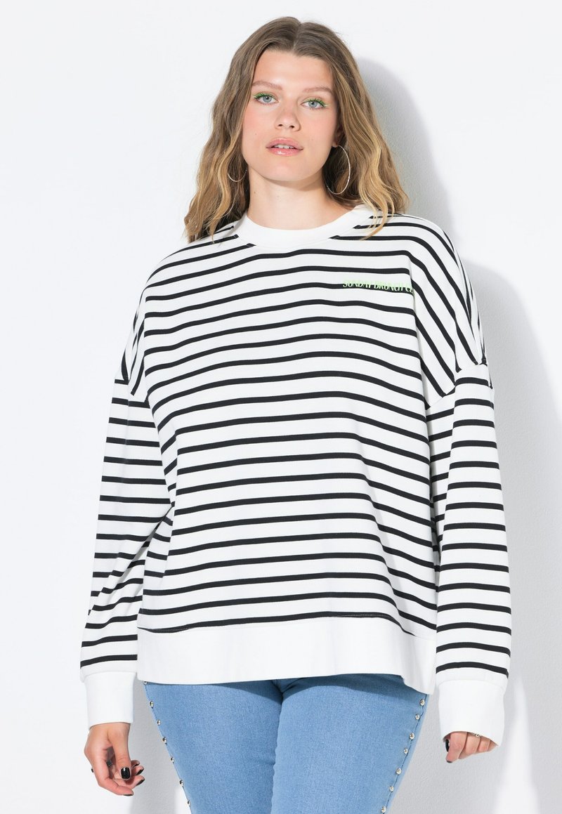 Sudadera a rayas en blanco y negro con mangas largas, que presenta un cuello redondo y un pequeño acento de logo verde en el pecho. Combinada con jeans azules.