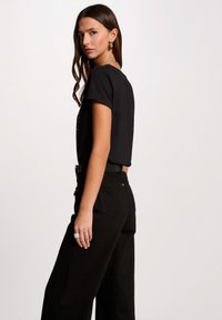 Maglietta nera a maniche corte, indossata con pantaloni neri a vita alta. Include un anello d'argento e presenta una consistenza liscia e morbida.