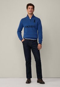 Suéter azul con cremallera en el cuello y logo "HACKETT" negro con el número 1, combinado con jeans de mezclilla oscuros y zapatos de cuero marrón. Fondo sencillo.
