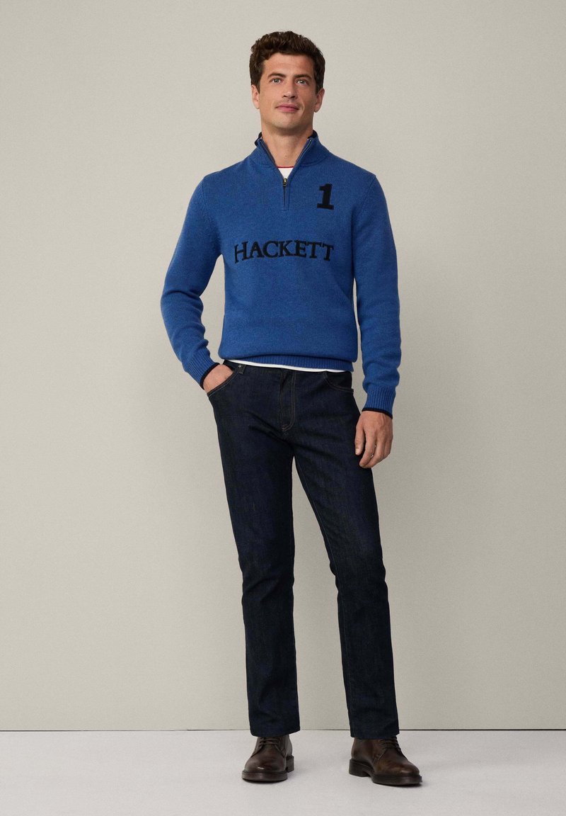 Pull à col zippé bleu avec le logo "HACKETT" noir et le numéro 1, associé à un jean en denim foncé et des chaussures en cuir marron. Fond uni.