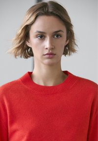 Röd stickad tröja med rund halsringning, med en slät textur och ribbad krage, bärs av en person med kort, ljusbrunt hår.