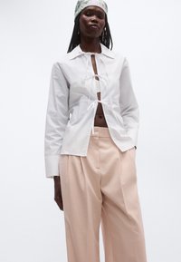 Fehér ing, melyet a mellrészen kötött megoldás díszít, hosszú ujjú és bő szabású, világos rózsaszín, magasderekú, bő szárú nadrággal párosítva.