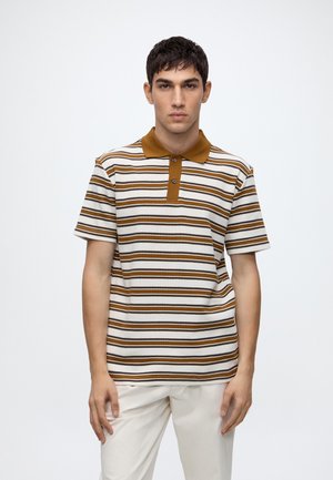Junger Mann mit kurzen dunklen Haaren, der ein braun-weiß gestreiftes Poloshirt und weiße Hosen trägt, steht vor einem schlichten Hintergrund.