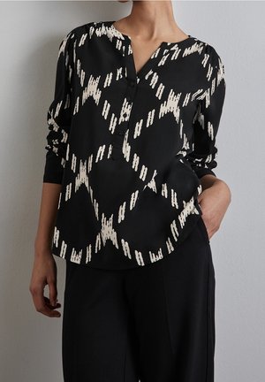 Vrouw die een zwarte blouse met lange mouwen draagt, voorzien van een wit abstract sterrenpatroon, en zwarte broek, staand met één hand in de zak.