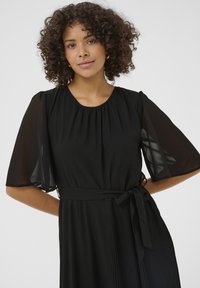 Rochie neagră cu un top din material neted, fustă pliată, mâneci puff din material transparent și o curea asortată în talie.