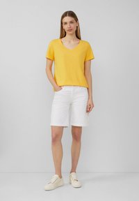 Gelbes V-Ausschnitt-T-Shirt, lockerer Schnitt, kombiniert mit weißen Shorts mit umgeschlagenem Saum. Das Model trägt hellfarbene Sneakers und steht vor einem schlichten Hintergrund.