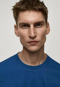 Modrá mikina s kulatým výstřihem s měkkou a hladkou texturou. Model má krátké, rozcuchané vlasy a je umístěn na neutrálním pozadí.