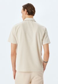 Camicia a maniche corte di colore beige chiaro con sottili righe verticali. Presenta un colletto a punte e una vestibilità comoda, realizzata in un tessuto testurizzato.