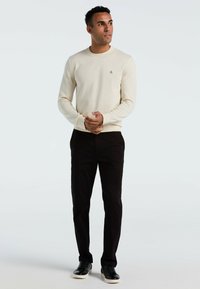 Licht beige ribbeltrui met een ronde hals en een klein logo, gecombineerd met zwarte broek en zwarte sneakers. Subtiele textuurdetails zichtbaar.