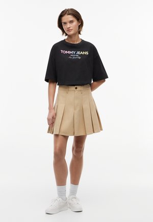 Jeune femme portant un t-shirt noir Tommy Jeans, une jupe plissée beige, des chaussettes blanches et des baskets blanches, debout devant un fond blanc.