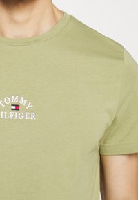 Camiseta de algodón verde oliva con cuello redondo y logotipo bordado de "Tommy Hilfiger" con un acento rectangular en rojo, blanco y azul.