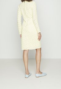 Kvinna som står med ryggen mot kameran iförd en knälång ärmlång gul spetsklänning och ljusblå platta skor mot en vit bakgrund.