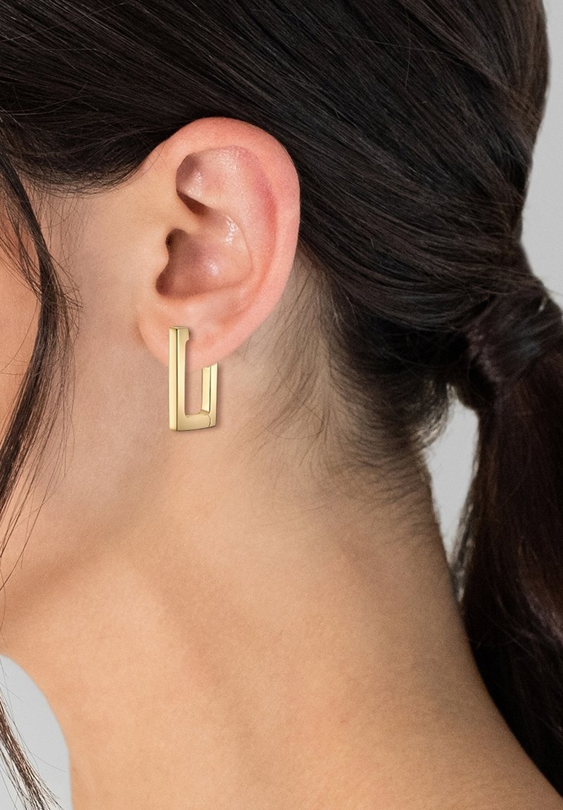 Auksiniai metaliniai auskarai geometrinės L formos, sklandus paviršius, mažo dydžio, uždėti ant ausies kaušelio, su tamsiais plaukais, sušukuotais atgal.