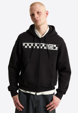 Junger Mann mit kurzen blonden Haaren, der einen schwarzen Vans Zip-Hoodie mit weißem Schachbrett-Logo trägt, steht mit den Händen in der Nähe der Taschen.