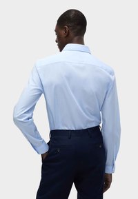 Camicia a maniche lunghe azzurro chiaro con colletto a punta, texture liscia e dettagli sulla parte posteriore, abbinata a pantaloni scuri con tasche laterali.
