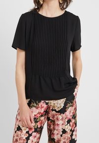 Blouse noire plissée à manches courtes et col rond; tissu léger; ourlet légèrement évasé; associée à un pantalon à imprimé floral.