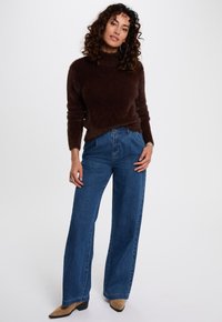 Maglione marrone peloso con colletto alto, abbinato a jeans in denim azzurri a gamba larga. Il modello indossa scarpe con tacco color beige e dettagli in gioielli d'argento.