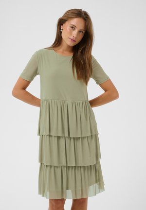 Jeune femme aux longs cheveux bruns portant une robe légère verte à étages avec des manches courtes, debout devant un fond uni.