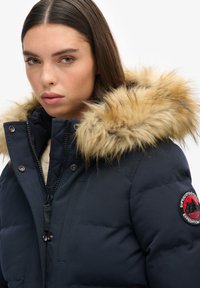 Veste en duvet marine avec une capuche doublée en fausse fourrure, dotée d'une fermeture éclair, de boutons-pression et d'un écusson logo rond sur l'épaule.
