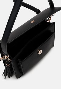 Sac à main en cuir noir avec une texture lisse, équipé d'un fermoir en ton doré, d'un compartiment intérieur et d'un pompon décoratif.