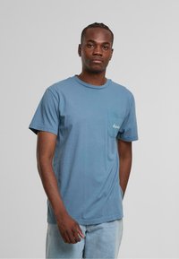 Blaues Baumwoll-T-Shirt mit Rundhalsausschnitt, kurzen Ärmeln und einer linken Brusttasche. Mit weißem, gesticktem Text auf der Tasche.