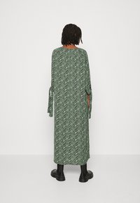 Langes grünes Kleid mit abstraktem schwarzen Muster, leichtes Material, weite Ärmel mit gebundenen Manschetten, knielange Länge, getragen mit schwarzen Stiefeletten.
