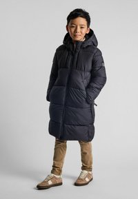 Manteau long noir rembourré avec une capuche, présentant des sections matelassées. Associé à un pantalon beige et des baskets marron avec des accents blancs.