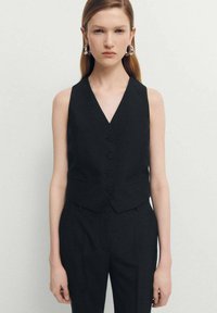 Čierny bezrukávový sveter s hlbokým výstrihom do V, piatimi gombíkmi a postrannými vreckami, vyrobený z textúrovanej látky, nosený cez zodpovedajúce nohavice.