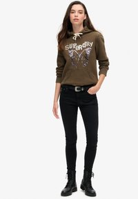 Olijfgroene hoodie met een vlinder sequindes ontwerp, drawstring capuchon en een kangoeroezak, gecombineerd met zwarte skinny jeans en zwarte combatboots.