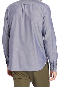 Uomo che indossa una camicia azzurro chiaro a maniche lunghe con bottoni e pantaloni verde oliva, visto da dietro, in piedi con le braccia rilassate.