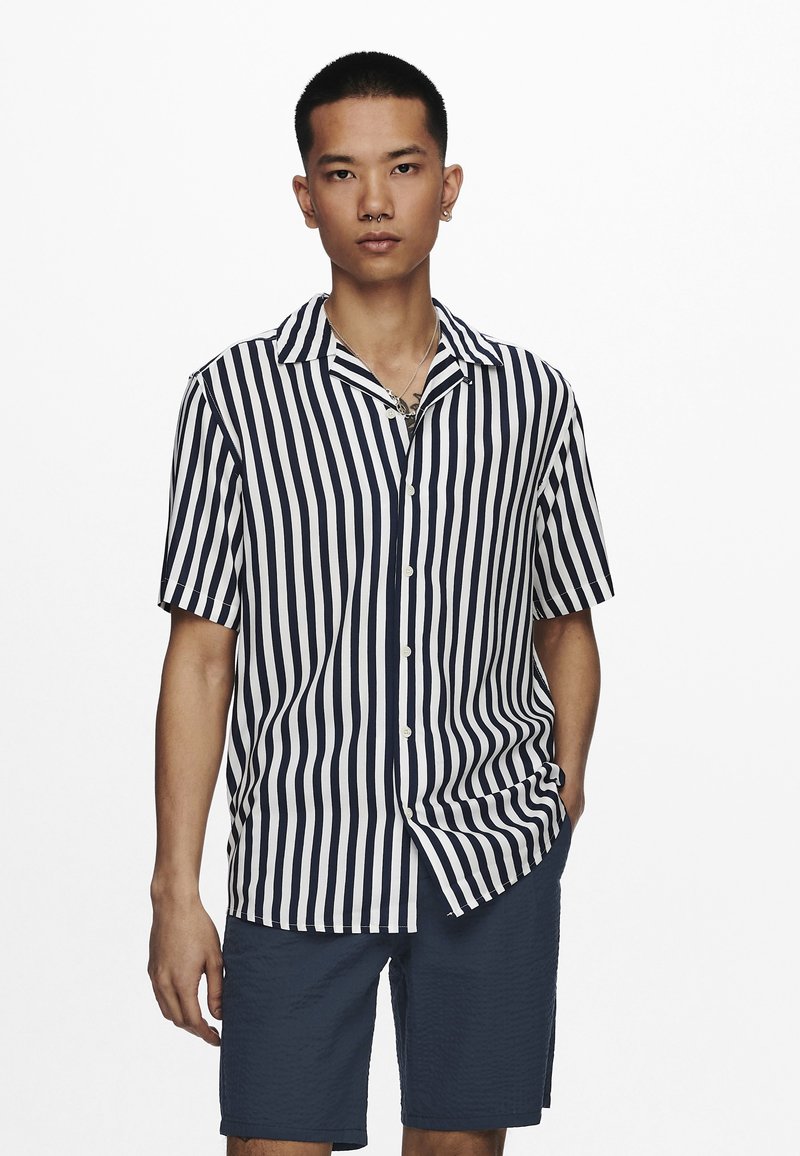 Korte mouw, knoopsluiting overhemd met marineblauwe en witte verticale strepen, een puntige kraag en een losse pasvorm. Gecombineerd met marineblauwe shorts.