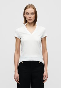 Άσπρο T-shirt με V-λαιμόκοψη και κοντά μανίκια, εφαρμοστή γραμμή, απαλό ύφασμα, με διακριτικές υφές στην αριστερή πλευρά του στήθους.