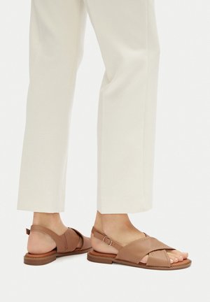 Sandali di pelle marrone con cinturini incrociati, chiusura con fibbia e suola piatta. Indossati con pantaloni bianchi a gamba larga.