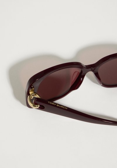 Tumedad punased Tory Burch'i prillid tumedate läätsede ja kuldse rõngastegaga templivarre detailidega, mis puhkusel valgel pinnal.