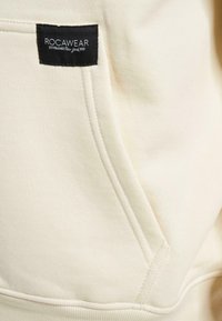 Hoodie color crema realizzato in morbido tessuto con tasca a marsupio e etichetta nera con il logo "ROCWARE" ricamato di lato.