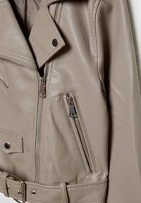 Beige faux leather motorcycle jacket with a notched collar, silver hardware, front zippers, and a belted waist. Smooth texture, fitted design.