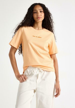 Kvinde med langt krøllet hår iført en ferskenfarvet 'Out of office'-t-shirt og hvide bindebukser, stående mod en ensfarvet hvid baggrund.