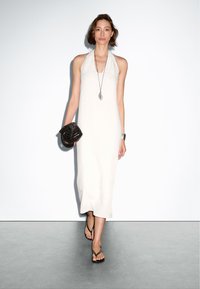 Langes, hellgraues Halterkleid mit gerader Silhouette. Eine strukturierte schwarze Handtasche und Flip-Flops runden den Look ab. Schlichter Halsreif als Akzent.