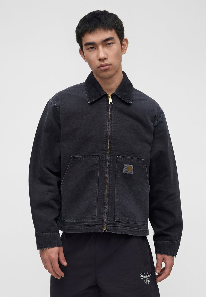 Junger Mann mit kurzem schwarzen Haar, trägt eine schwarze Reißverschlussjacke mit großen Fronttaschen und schwarze Hose vor einem einfarbigen Hintergrund.